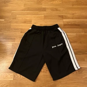 Palm Angels svarta shorts med vita ränder - Svarta shorts från Palm Angels med vita sidoremsor och broderad logga framtill. Modellen har elastisk midja med snörning och två fickor. Sportig vibe och loose fit, perfekta för chill eller träning. Materialet är mjuk polyester som känns skönt mot huden.