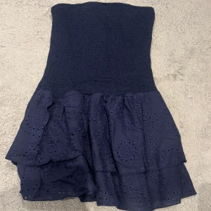 Marinblå axelbandslös kortklänning - Säljer en marinblå axelbandslös kortklänning med smockad överdel och volangkjol i broderad bomull. Kjolen har två lager med fina hålbroderier som ger en cool och feminin vibe. Perfekt för dig som gillar att sticka ut med detaljer och älskar sommarlooken.