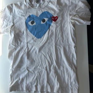 Vit Comme des Garçons Play t-shirt S - Snygg vit t-shirt från Comme des Garçons Play i storlek S. Framsidan har ett stort blått hjärta med ögon och ett mindre rött hjärta med ögon. Klassisk rund hals och korta ärmar.