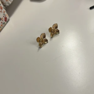 Guldiga blomformade örhängen - Supersöta örhängen i form av blommor med detaljerade kronblad och små pärlor i mitten. De är i guldig färg och har en elegant, feminin design som verkligen poppar. Perfekta för att ge din look en extra touch av stil.