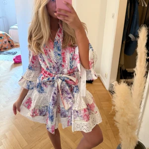 Blommig vit kortklänning från Bella barnet - Superfin vit kortklänning från Bella barnet med blommigt mönster i rosa, lila och blå toner. Klänningen har volangdetaljer, spets och knytband i midjan. Trekvartsärmar med volang och omlottkänsla. (Finns ej kvar)