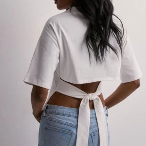 Nelly tie back topp - Superfräsch vit croppad topp i bomull med breda korta ärmar och snygg knytning i ryggen. Toppen har en tight passform runt midjan och en öppen rygg som ger en cool och trendig look. Perfekt att styla med jeans eller kjol för en modern vibe.