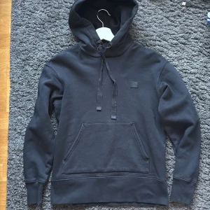 Svart hoodie från Acne Studios - Säljer en svart hoodie från Acne Studios med klassisk smiley-patch på bröstet. Tröjan har huva med snörning, stor magficka och långa ärmar. Mjuk och skön bomullskvalitet, perfekt för chill dagar. Passar dig som gillar stilren och enkel design.