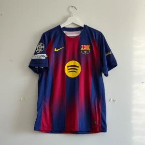 FC Barcelona Lamine Yamal #10 matchtröja - Säljer en  FC Barcelona matchtröja med Lamine Yamal #10 på ryggen. Tröjan har klassiska blå och röda ränder, gula detaljer och klubbmärke på bröstet. Materialet är lätt och ventilerande polyester, med Champions League-patch och Spotify-logga på framsidan.