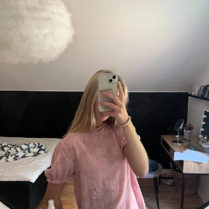 Söt rosa blus med knyt i nacken - En ljusrosa blus med blommigt mönster och korta puffärmar. Blusen har en fin knytning i nacken och volangdetaljer vid ärmsluten. Perfekt för dig som gillar en romantisk och söt stil. Osäker var den är ifrån..