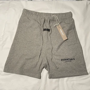 Essentials shorts - Snygga grå shorts från Fear of God Essentials med broderad logga på benet. De har elastisk midja med snörning och är gjorda i mjuk bomull, perfekta för chill dagar. Klassisk passform och stilren look som funkar till allt.