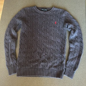 Mörkblå kabelstickad tröja Ralph Lauren - Säljer en mörkblå kabelstickad tröja från Ralph Lauren i storlek XS. Tröjan har rund hals, ribbade muddar och ett litet rött broderat Polo-logo på bröstet. Perfekt för dig som gillar klassisk stil med snygga detaljer och mjukt material.