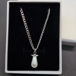 Silverhalsband med pärlhänge - Stilrent halsband i silverfärgad metall med en kedja och ett droppformat vitt pärlhänge. Hänget har en liten detalj upptill som ger det en unik look. Perfekt för dig som gillar minimalistiska smycken med en twist.