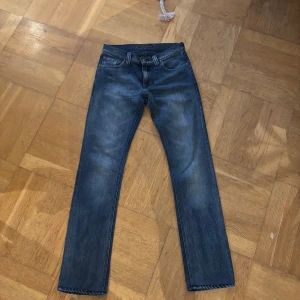 Mörkblå Nudie Jeans - Snygga mörkblå jeans från Nudie Jeans med klassisk femficksdesign. Jeansen har feta slitningar och snygg tvätt, samt Nudie-logga på bakfickan. Perfekt för dig som gillar en tidlös och avslappnad stil.