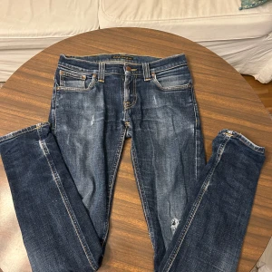 Blå jeans från Nudie Jeans W27 L32 - Snygga blå jeans från Nudie Jeans med klassisk femficksdesign och coola slitningar. De har kontrastsömmar i gult och Nudie Jeans-logga på bakfickan. Passformen är rak och materialet är slitstarkt denim. Perfekt för dig som gillar streetstyle och vill ha ett par jeans med attityd.