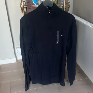 Svart polotröja Tommy Hilfiger L - Svart polotröja från Tommy Hilfiger i storlek L. Tröjan har en half zip-dragkedja vid halsen och diskret Hilfiger-logga på bröstet. Långärmad och tillverkad i mjuk bomull, perfekt för dig som gillar stilrena och klassiska plagg.