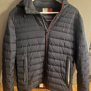 Blå pufferjacka från Tommy Hilfiger - Snygg blå pufferjacka från Tommy Hilfiger med huva. Jackan har dragkedja framtill, två fickor med dragkedja och diskret logga på bröstet. Perfekt för kyliga och höstiga dagar med stil