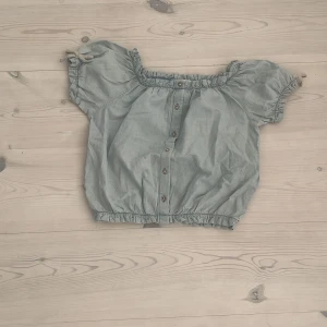 Grön blus med knappar från Zara Kids - Söt ljusgrön blus från Zara Kids med korta puffärmar och resår vid hals, ärmslut och nederkant. Blusen har knappar framtill och små knytband på ärmarna. Perfekt för en avslappnad och trendig look under varma dagar.