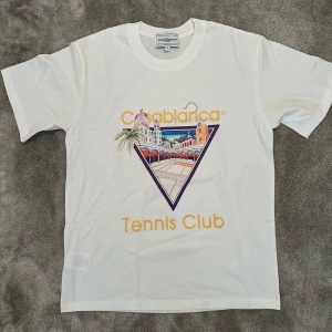 Vit Casablanca Tennis Club t-shirt S - ✨ Casablanca T-shirt ✨ Vit t-shirt från Casablanca 🤍 med färgglad grafik och texten “Tennis Club” på framsidan 🎾. 	•	👕 Klassisk rund hals och korta ärmar 	•	🧵 Tillverkad i mjuk bomull med detaljer i viskos och silke för en lyxig känsla 	•	🌟 Perfekt för dig som gillar sportig och trendig stil  En t-shirt som kombinerar komfort, lyx och modern design.