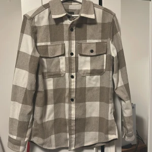 Beige och vit rutig overshirt - Snygg overshirt i beige och vitt rutigt mönster med klassisk krage och svarta knappar. Två bröstfickor med lock och knapp, samt långa ärmar med knapp vid mudden. Perfekt lager-på-lager-plagg för en chill och trendig look.
