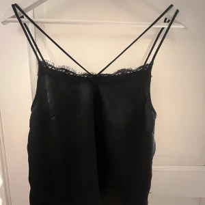 Svart linne Gina Tricot - Säljer ett svart linne från Gina Tricot i glansig polyester med tunna axelband och spetsdetaljer vid halsringningen. Linnet har en slinky och elegant look med figurnära passform och ärmlös design. Perfekt för dig som gillar minimalistisk stil med en touch av lyx.