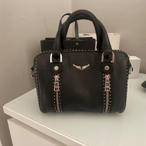 Svart handväska Zadig & Voltaire - Cool svart handväska från Zadig & Voltaire i skinn med silverfärgade detaljer och dubbla dragkedjor framtill. Väskan har små nitar längs kanterna och ett metallmärke med vingar på framsidan. Perfekt storlek för det viktigaste och har både handtag och axelrem. Ny priset ligger på ca 4000kr💞