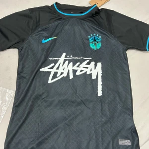 Nike x BRAZIL svart fotbollströja Stüssy - Snygg svart fotbollströja från Nike i samarbete med CBF och Stüssy. Tröjan har korta ärmar, turkosa detaljer och CBF-emblem på bröstet. Stüssy-logga i vitt framtill. Tillverkad i lätt och ventilerande material, perfekt för träning eller streetwear.
