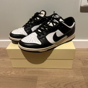 Nike Dunk Low svart/vit sneakers - Nike Dunk Low sneakers i svart och vitt med klassisk Swoosh på sidan. Skorna har snörning, perforerad tå och platt sula. Tillverkade i skinn med låg siluett och rund tå. Perfekt för dig som gillar stilrena och tidlösa sneakers.