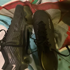 Nike Air Zoom svarta fotbollsskor - Säljer ett par svarta Nike Air Zoom fotbollsskor med snyggt mönstrad ovandel och klassisk swoosh på sidan. Skorna har snörning och platt sula med dobbar för gräsplan. Materialet är syntet och designen är riktigt sportig och modern.