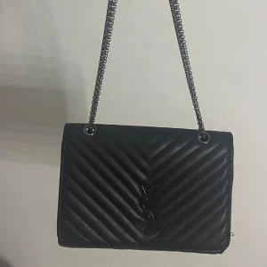 Svart axelväska från YSL - Säljer en svart axelväska från YSL med kedjerem och ikonisk YSL-logga framtill. Väskan är i skinn och har ett snyggt chevronmönster. Insidan har dragkedjefack och magnetlås. Tveka inte att skriva vid funderingar 💕(Köpt utomlands)