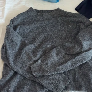 Grå stickad tröja med rund hals - Mysig grå stickad tröja med rundad hals och långa ärmar. Perfekt för kyliga dagar och enkel att matcha med jeans eller mjukisbyxor. Tröjan har en klassisk, loose passform och ribbade muddar vid hals, ärmslut och nederkant.