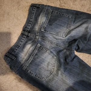 Blå bootcut jeans med slitningar - Säljer ett par blå jeans med coola slitningar och tvättade detaljer. Modellen har bootcut-ben och klassiska bakfickor. Jeansen har normal passform och är tillverkade i jeansmaterial. Innerbenslängd 80cm, midjemått ≈ 38cm