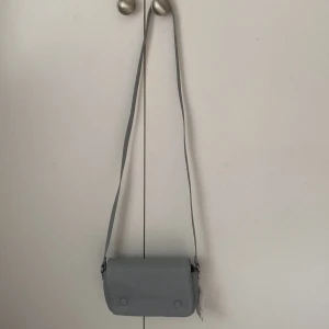 Blågrå axelväska från H&M - Snygg blågrå axelväska från H&M med stilren design och mjukt syntetmaterial. Väskan har ett lock med två dekorativa knappar framtill. Insidan är beige. ALDRIG ANDVÄND PRISLAPP ÄR KVAR!