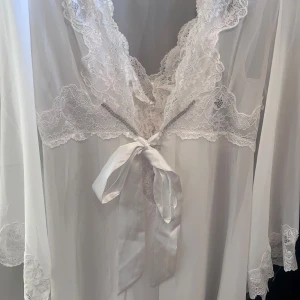 Vit kort spetsklänning med omlott - Supersöt vit kort klänning i satin med spetsdetaljer och omlottmodell. Klänningen har långa vida ärmar med spets vid ärmslut, v-ringad hals och ett brett satinband att knyta i midjan. Perfekt för dig som gillar romantisk och feminin stil.