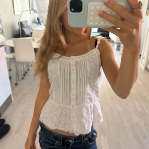 Vit spetslinnetopp från Superdry - Supersöt vit linnetopp från Superdry med tunna axelband, spetsdetaljer och knappar framtill. Toppen har romantiska volanger och broderade partier som ger en bohemisk känsla. Perfekt för varma dagar och somriga outfits.
