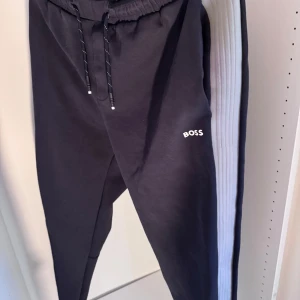 Svarta mjukisbyxor från BOSS - Svarta mjukisbyxor från BOSS med vit logga på låret och breda ribbade sidopaneler i ljusgrått. Byxorna har elastisk midja med snörning och en sportig look, perfekta för chill eller träning.