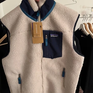  Beige fleeceväst från Patagonia - Säljer en beige fleeceväst från Patagonia med marinblå detaljer och dragkedjor. Helt ny med prislapp kvar! Hör av er vid funderingar 