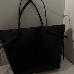 Svart shoppingväska i skinn - Stor och stilren svart shoppingväska med minimalistisk design och två långa handtag. Väskan har snygga knyt-detaljer på sidorna och är tillverkad i slätt skinnmaterial. Perfekt för dig som vill ha plats för allt och ändå se clean ut.