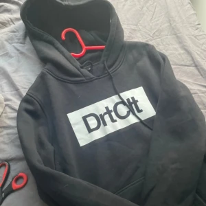 Svart hoodie DrtCtt XL - Svart hoodie från DrtCtt i storlek XL med stor vit logga på bröstet. Klassisk huva med snörning och mjukt material som känns skönt mot huden. Perfekt för chill dagar eller när du vill ha en clean streetwear-look.