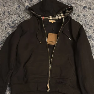 Svart Burberry hoodie med dragkedja XL - Svart hoodie från Burberry i storlek XL med klassiskt rutigt foder i huvan och insidan. Tröjan har huva med snörning, två fickor fram och dragkedja. Materialet är mjuk bomull och polyesterblandning. Perfekt för dig som vill ha en stilren och lyxig look.