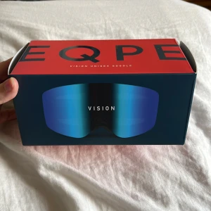 EQPE Vision blå skidglasögon - Säljer ett par EQPE Vision skidglasögon med spegelblå lins och svart ram. Glasögonen har en bred, rundad form och ett elastiskt svart band med vit EQPE-logga. Perfekt för dig som vill ha cool stil i backen och bra skydd mot snö och sol. Dem är använd en vinter men köpt nya som jag gillar mer inget fel på dem.