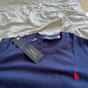 Mörkblå t-shirt från Polo Ralph Lauren - Snygg mörkblå t-shirt från Polo Ralph Lauren med klassisk röd logobrodering på bröstet. Modellen är kortärmad och har en rund halsringning. Materialet är mjuk bomull som känns skönt mot huden. Perfekt för dig som gillar stilrena och tidlösa plagg.