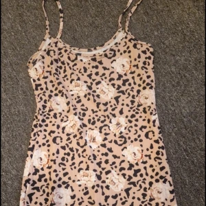 Beige leopardmönstrad klänning med blommor - Säljer en beige kortklänning med leopardmönster och stora blommor i vitt och ljusrosa. Klänningen har smala axelband och är ärmlös, med en rak och slim passform. Perfekt för dig som gillar att sticka ut med coola mönster och mjuka färger.