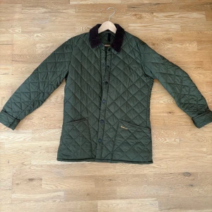 Grön quiltad Barbour jacka M - Säljer en grön quiltad jacka från Barbour med klassisk brun manchesterkrage. Jackan har tryckknappar framtill, två stora fickor och Barbour-logga broderad på ena fickan. Perfekt för dig som gillar stilren och tidlös design. It has only been used for one fall by me - so approximately it has 3 months of use. This leaves the jacket in a good as new condition for its new owner.There is no one who smokes in my house and we do not have any pets. 
