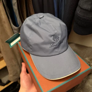 Loro Piana keps  - Snygg blå keps från Loro Piana med broderad logga framtill. Kepsen har en klassisk form med böjd skärm i vit och insida med rutigt mönster. Tillverkad i polyester och bomull, perfekt för dig som vill ha en stilren och exklusiv look.
