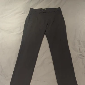 Slim grå kostymbyxor Jack & Jones - Snygga grå kostymbyxor från Jack & Jones Pants Studio med slim passform. Byxorna har klassiska fickor fram och bak, bälteshällor och stängs med knapp och dragkedja. Perfekt för dig som gillar stilrena och moderna byxor med en smal siluett.