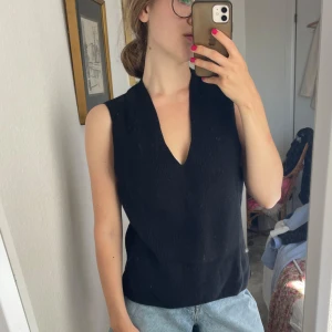 Svart stickad ärmlös topp med v-ringning - Svart stickad topp med djup v-ringning och ärmlös design. Modellen är rak och har små slitsar i sidorna för en chill look. Perfekt att styla med jeans eller shorts för en enkel men snygg outfit.