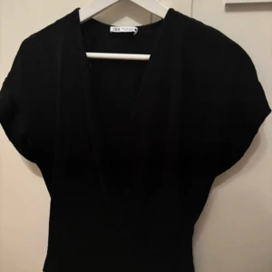 Svart omlott blus från Zara - Snygg svart blus från Zara med omlott-design och v-ringad front. Blusen har korta ärmar och en stilren, figurnära passform som ger en modern look. ALDRIG ANVÄND, ENDAST TESTAD!!!!