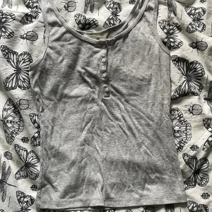 Grå ribbad linnetopp från H&M - Säljer en grå ribbad linnetopp från H&M med rund halsringning och knappar framtill. Toppen är ärmlös och har en slim passform som sitter snyggt på kroppen. Perfekt att styla med jeans eller kjol för en enkel och clean look.