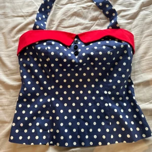 Prickig blå korsett med röd krage - Snygg korsett-topp i marinblått med vita prickar och en cool röd krage. Toppen har breda axelband, dekorativa svarta knappar framtill och en figurnära passform. Perfekt för dig som gillar retrovibbar och vill sticka ut med färg och mönster.