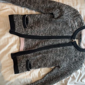 Cardigan/kofta - Säljer en snygg gråmelerad stickad kofta från By Malene Birger med svarta kanter och rund hals. Koftan har långa ärmar och två fickor framtill. Perfekt för lager-på-lager och chill stil.