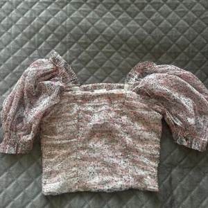 Blommig blus med puffärmar - Säljer en söt blommig blus med puffiga korta ärmar och fyrkantig halsringning. Toppen har ett ljust mönster i rosa, vit och grå med små blommor och smock vid ärmslut. 