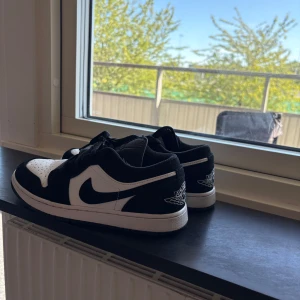 Nike Jordan 1 Low svart/vit strl 41 - Nike Jordan 1 Low i svart och vitt med klassisk swoosh på sidan och Air Jordan-logga på hälen. Skorna har snörning, rund tå och platt sula. Ovandelen är i skinn och syntet, vilket ger en clean och sportig vibe. Perfekt för dig som gillar streetstyle och sneakers med ikonisk design.