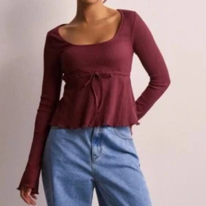 Vinröd ribbad topp  - Säljer en vinröd långärmad topp från Nelly. Den har ett knytband i midjan som ger en snygg siluett. Toppen har lätt utsvängd nederkant och lite vidare ärmar 