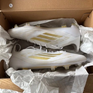 Adidas F50 Elite - Säljer ett par Adidas F50 fotbollsskor i vitt med guldiga detaljer och klassiska tre ränder på sidan. Skorna har snörning, platt sula med dobbar och är tillverkade i syntetmaterial för lätt känsla på planen. Perfekt för dig som vill sticka ut med stil på matchen. Säljer dessa för 1699 nypris 3100